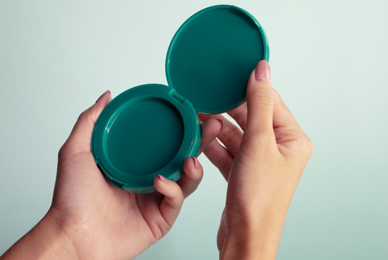 A pair of hands holding open a teal compact mirror made entirely of Eastman Cristal One Renew. 