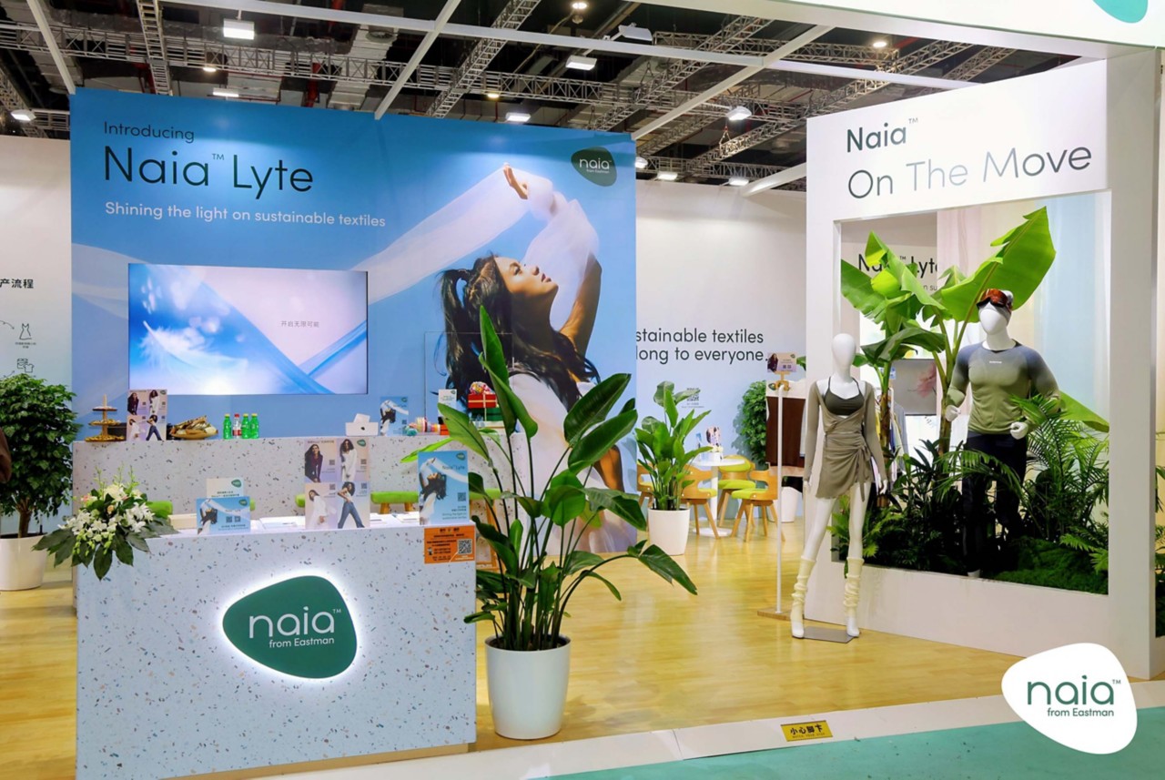 The booth displays mannequins wearing garments made with Naia Lyte and various plants. 