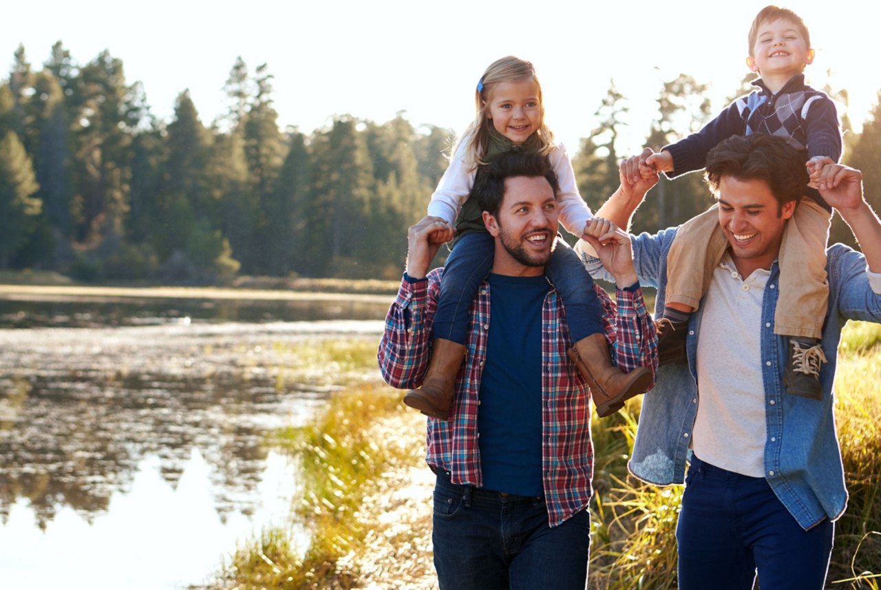 Two men walking by lake with children on shoulders. 