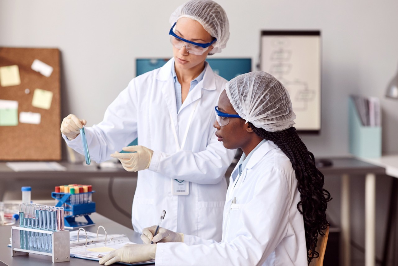 Two women in lab coats and protective gear looking at test tube in a lab setting 