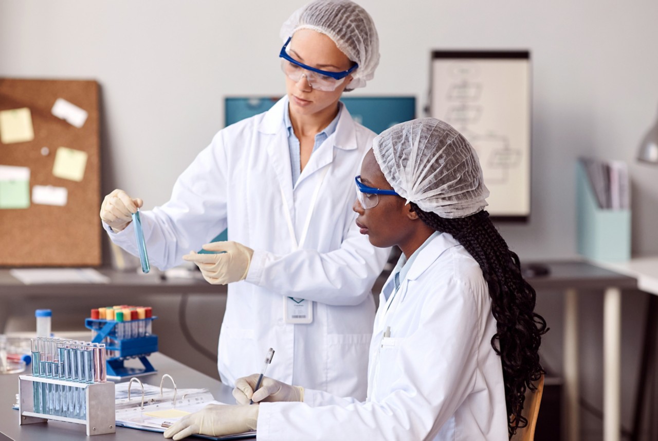 Two women in lab coats and protective gear looking at test tube in a lab setting. 